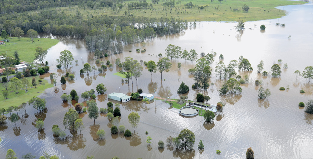 A rural property is flooded, with several buildings partially submerged. There is some dry land in the background of the photo.