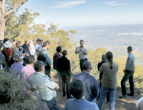 A photo of several people standing on hill overlooking the landscape. A person is standing in front of the main group talking and gesturing with his hands.