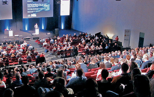 An overhead image of the conference presentation stage. There are two levels of attendees listening to the main speaker on the front stage.