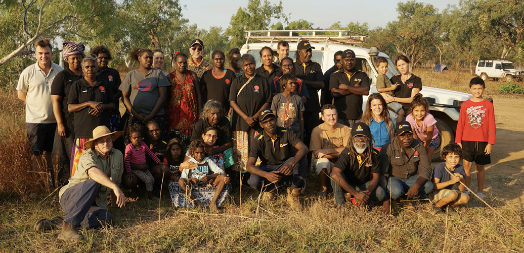 A photo of the groups, taken outside, with a dirt road and parked vehicles in the background.