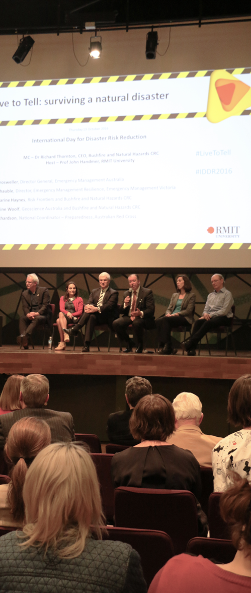 A photo of the ‘Live to tell’ forum. A panel of speakers is on stage and a slide about ‘Live to tell: surviving a natural disaster’ is on the screen behind them. Members of the audience are looking at the stage.