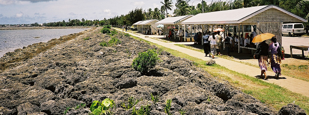 A photo of Nuku’alofa, the capital of the Kingdom of Tonga, showing the sea front and rocky shore with bus shelters and people directly behind them.