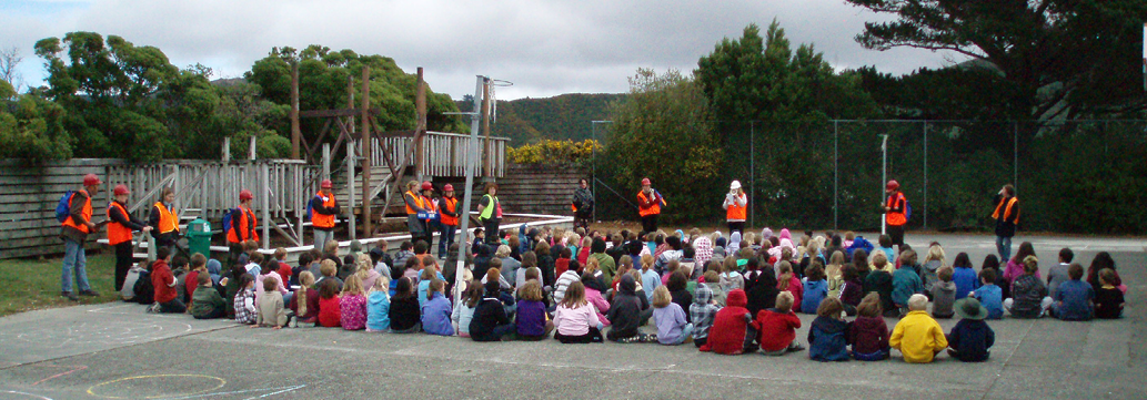 A photo of students and staff receiving hazards education and emergency response training from emergency workers. The students and staff are sitting on the ground in an outdoor area, with emergency staff around them. 