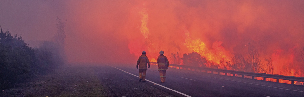 Photo of two firefighters walking down a road towards a smoky bushfire that is burning on one side of the road.