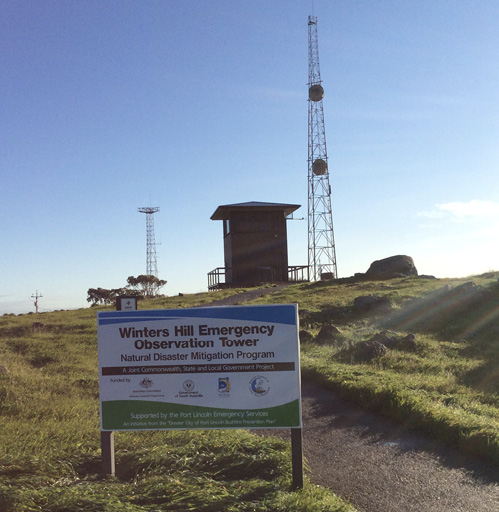 Image of the observation tower, which is sitting on grassland. In front is a sign stating that it is the Winters Hill Emergency Observation Tower.